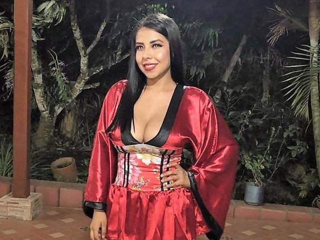SamanthaKlaus with long black hair, red silk dress, black trim, low neckline showing her big tits, smiling, standing outside, night, forest background, potted plant on tiled floor.