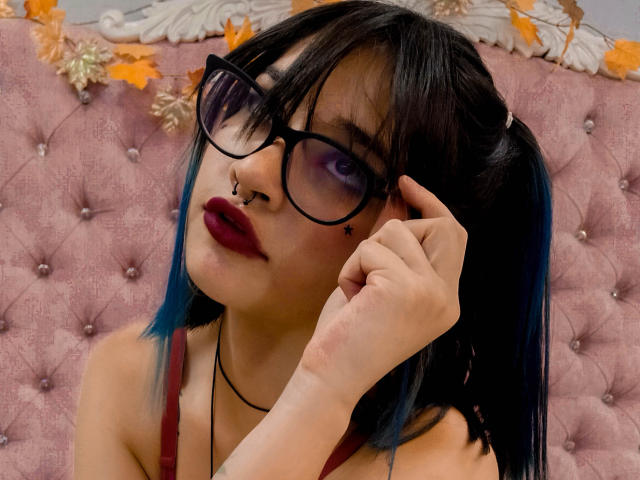 CameroonSweet, met donkerblauw haar, zwarte bril, neusring en rode lippenstift, die haar haar aanpast en een lichte glimlach toont, draagt een rode top en zit op een roze gestoffeerde stoel..