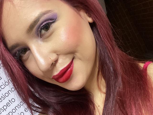 ValeryScot with red lipstick, purple eyeshadow, nose ring, and pink hair, smiling at the camera. ValeryScot's face is slightly tilted, showing her shiny skin and cute smile.