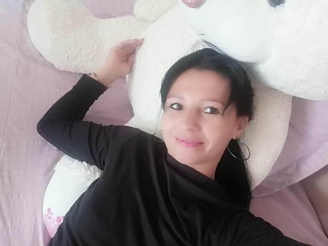 KratosYSara, avec des cheveux noirs, portant un haut noir, allongée sur un ours en peluche géant blanc, souriante, avec des boucles d'oreilles pendantes, du rouge à lèvres rose, des seins de taille moyenne, un drap de lit rose..