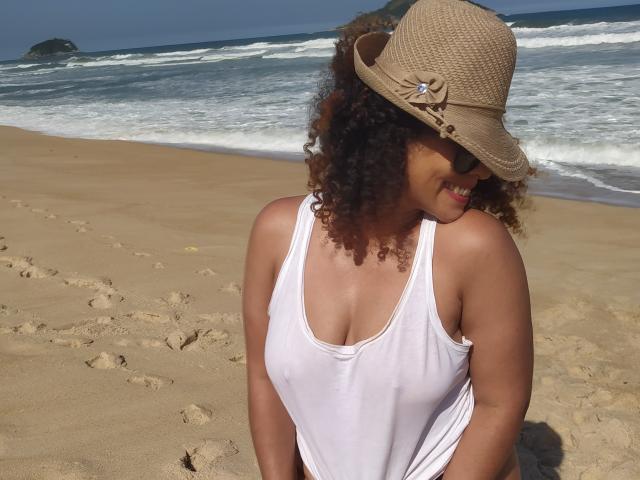 Curly-haired VolupMilf with tan skin wearing a beige straw hat and white tank top, showing small tits, stands on a sandy beach with waves in the background.