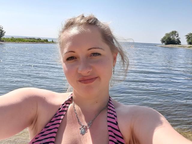 Samiliya, de piel clara, con cabello rubio recogido en una coleta, vestida con un top de bikini a rayas rosa y negro, mostrando sus pechos, de pie junto a un lago, sonriendo a la cámara..