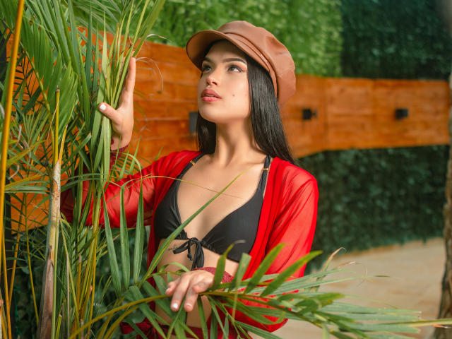April Novoa, con cabello negro largo, gorra marrón, top rojo transparente, bikini negro, sosteniendo hojas de palma, mirando hacia arriba con la boca ligeramente abierta, mostrando sus pechos..