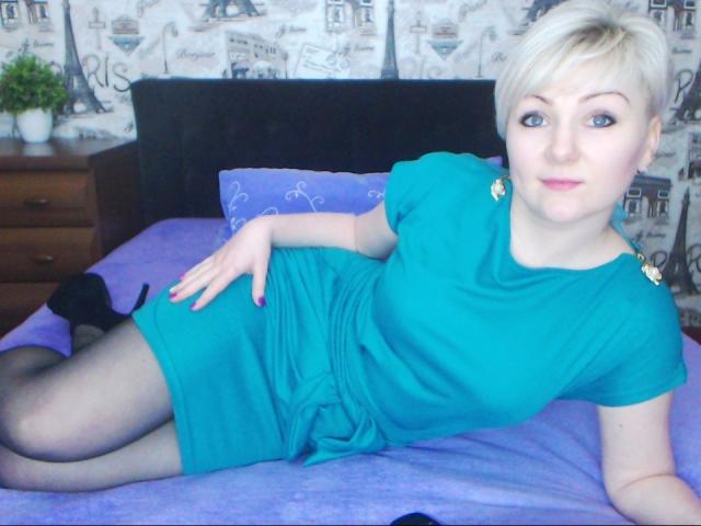 BlondPearX, avec des cheveux blonds courts, une robe verte, des talons hauts noirs et des bas noirs, allongée sur un lit violet, une main sur ses fesses, regardant l'appareil photo..