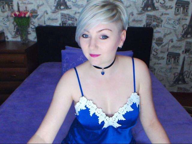 BlondPearlX con capelli biondi corti, vestito azzurro con pizzo, rossetto rosa, collana nera e ciondolo azzurro, seduta su un letto viola, che mostra piccoli seni e pelle chiara..