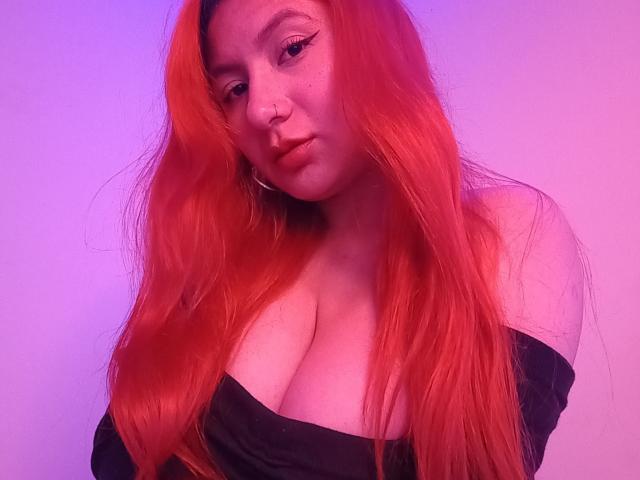 AradiaX with red hair, nose ring, and black off-shoulder top, showing her big tits against purple lighting. She has a pouty expression and long, wavy hair.
