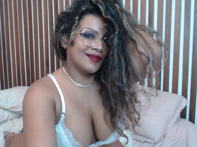 EbonyRose69 with curly brown hair, glasses, red lipstick, white lace bra, gold chain, smiling, showing big tits, sitting on bed, hand in hair.
