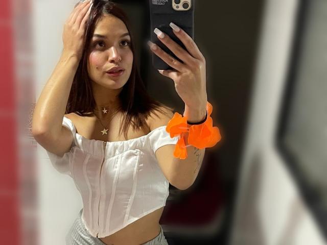 AprilNovoa with brown hair, wearing a white off-shoulder crop top and grey pants, holding a phone, orange scrunchie on wrist, star necklace, pouting, mirror selfie, blushing.