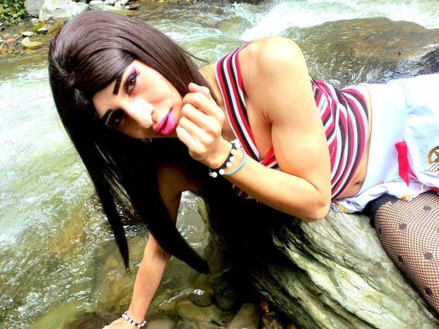 Catalina Castro, avec de longs cheveux bruns, du rouge à lèvres rose, un débardeur rayé, une jupe blanche, des bas résille, appuyée sur un rocher dans un ruisseau, la main près de ses lèvres roses, des bracelets au poignet..