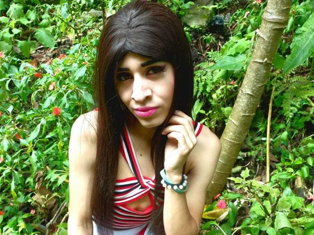 Catalina Castro mit langem, dunklem Haar, rosafarbenem Lippenstift und einem roten Streifenkleid, die ihre Wange berührt, ein grüner Armreif, ein Wald im Hintergrund, die in die Kamera schaut, sexy..