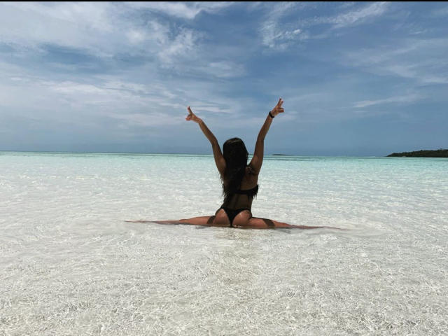 StormyKiss, mit dem Rücken zur Kamera, sitzt in flachem, klarem Wasser, die Arme erhoben, trägt einen schwarzen String-Bikini, hat langes, dunkles Haar, im Hintergrund ist ein blauer Himmel und ein ruhiges Meer zu sehen..