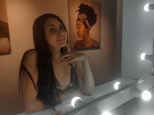 AmbeerBlack, avec ses longs cheveux noirs, est assise devant un miroir éclairé, souriante, montrant sa poitrine, une main près de sa bouche, mur brun..