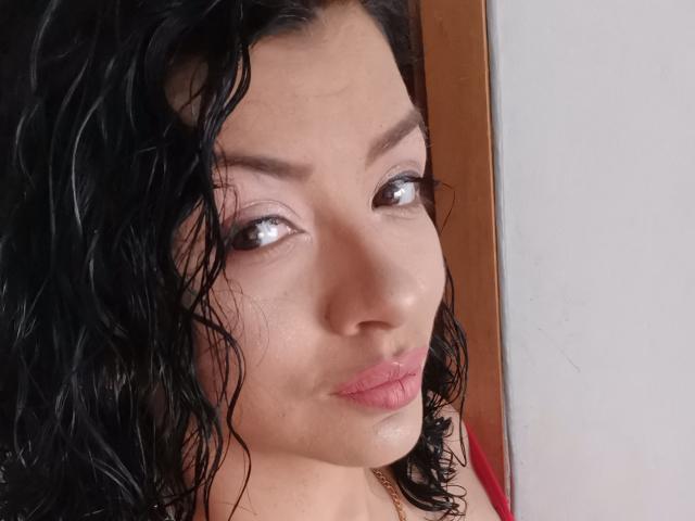 IsabellaLnd tem cabelos pretos encaracolados e úmidos, olhos castanho-claros e batom rosa. Seu rosto mostra um leve franzido. Ela está vestindo uma blusa vermelha e um colar dourado..