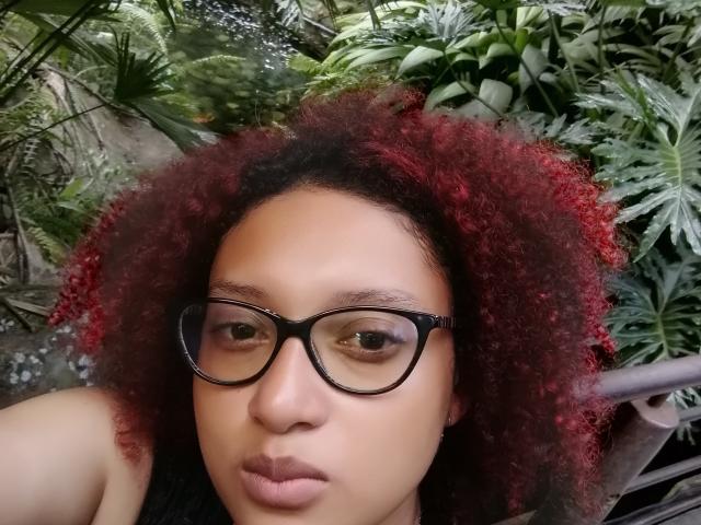 VioletaSwitch has dark curly red hair, black glasses, and a serious expression. She's looking at the camera with her brown eyes and slightly pouty lips.