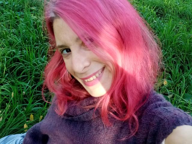ASexyLady with pink hair, smiling, wearing a purple knit turtleneck, grass in the background, looking at the camera, showing her teeth, cute, and attractive.