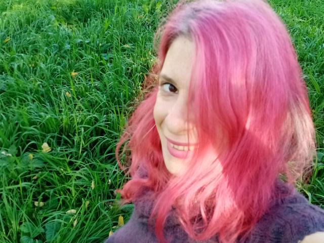 Een aantrekkelijke vrouw met roze haar, die lacht, draagt een donkerpaarse, pluizige trui, heeft een lichte huid en staat in het groene gras. Haar borsten zijn niet zichtbaar, alleen haar schattige gezicht..