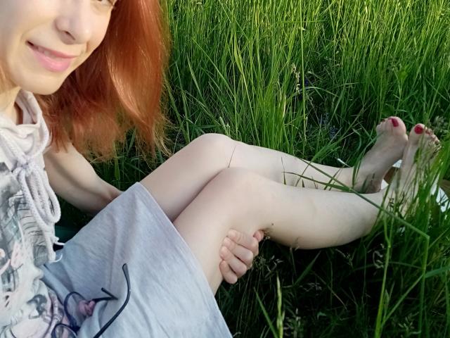 ASexyLady with red hair, pale skin, and red toenails, sitting in grass, legs crossed, wearing a gray dress, smiling, showing her bare legs and feet, selfie.