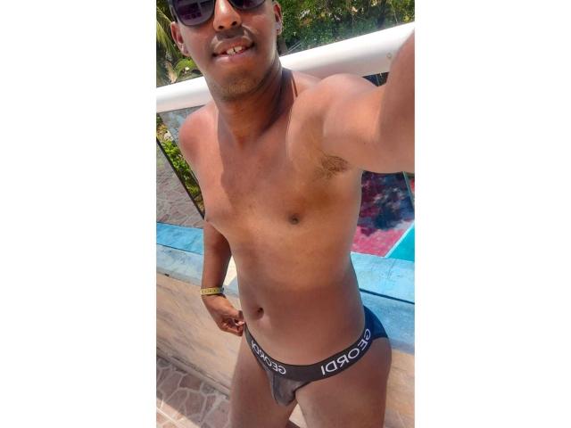MrJhon69, dunkelhäutig, trägt einen schwarzen Speedo, zeigt einen schlaffen, kaum sichtbaren Penis, mit einer goldenen Armbanduhr und schwarzen Sonnenbrille, lächelt im Freien..