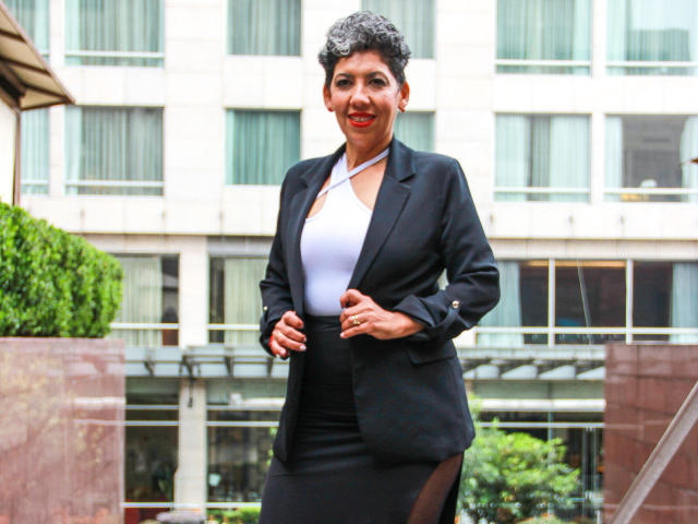 JuliaNewell with short curly gray hair, wearing a black blazer, white halter top, and black skirt, standing outdoors, smiling, hands on jacket, showing her small ass.