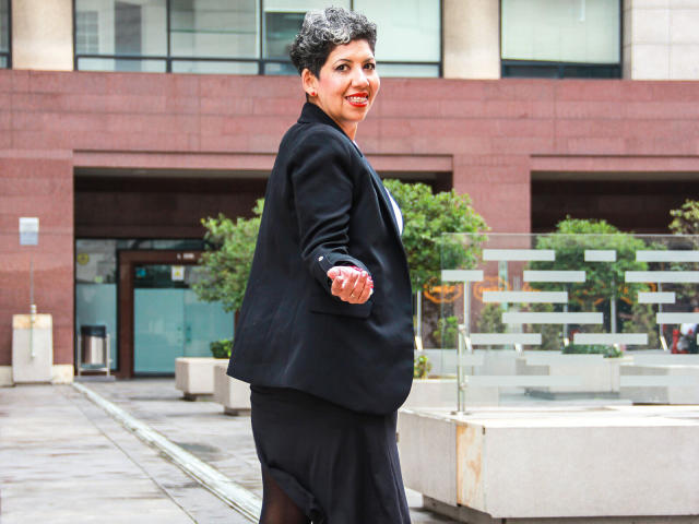 JuliaNewell with short curly black hair, wearing a black suit, standing outside, smiling, looking over her shoulder, red hand covering her crotch, urban background, daytime, confident.