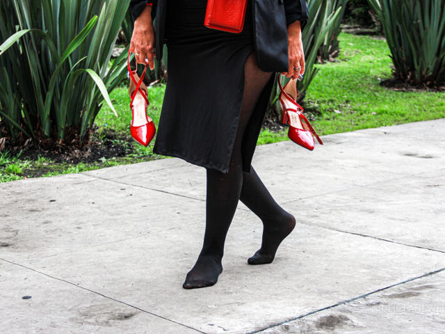 JuliaNewell walks on a concrete sidewalk, wearing black sheer stockings, a black skirt, holding red high-heeled shoes in both hands, and a red purse.