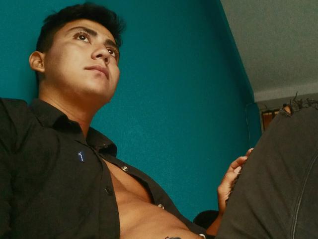 DorkHot, com cabelo preto curto, olhando para cima, vestindo uma camisa preta aberta, revelando um peito musculoso e sem camisa, e segurando suas calças pretas. Parede azul-turquesa ao fundo..