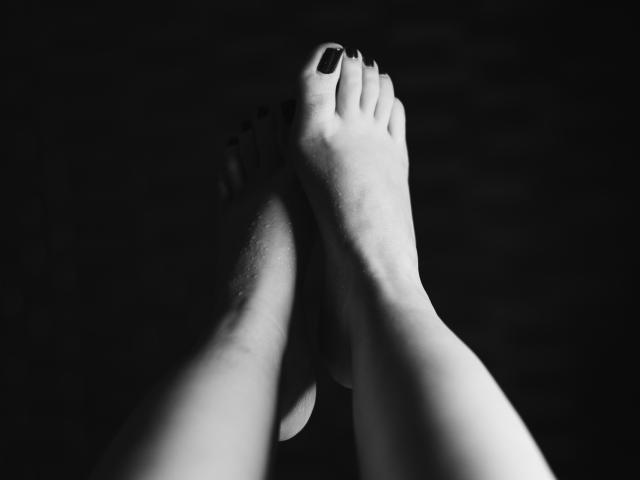 I piedi nudi di AbbyToriner, con le unghie dei piedi dipinte di nero, le dita rivolte verso l'alto, leggermente divaricate, che mostrano una sottile ombra sulla pianta del piede. Immagine scura, ad alto contrasto..