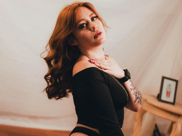 Abby Toriner mit roten, gewellten Haaren, einem schwarzen Top mit einem Schulter frei, und schwarzer Unterwäsche, die über die Schulter schaut, eine Hand am Hals, ein Nasenring, ein Tattoo am Arm, eine Armbanduhr, rote Nägel, sexy..