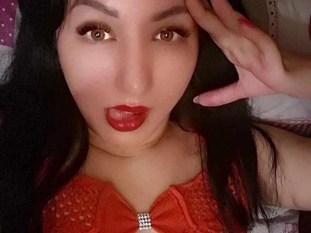 HotBestia con lápiz labial rojo y cabello oscuro, lengua afuera, haciendo un gesto de la paz, vistiendo una blusa de encaje naranja, tiene ojos marrones y una cara linda, está acostada..