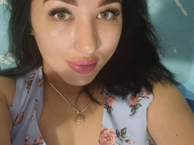 A HotBestia tem pele marrom clara, sobrancelhas escuras, olhos verdes, batom rosa, cabelo preto, um vestido floral, um colar de ouro com um pingente de rosa, e está com uma expressão fofa, ligeiramente emburrada..