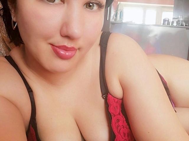 HotBestia, avec ses lèvres roses, ses yeux marron et son soutien-gorge en dentelle noir et rouge, montre ses seins volumineux et ses fesses douces dans une chambre. Sa peau est claire et elle sourit légèrement..