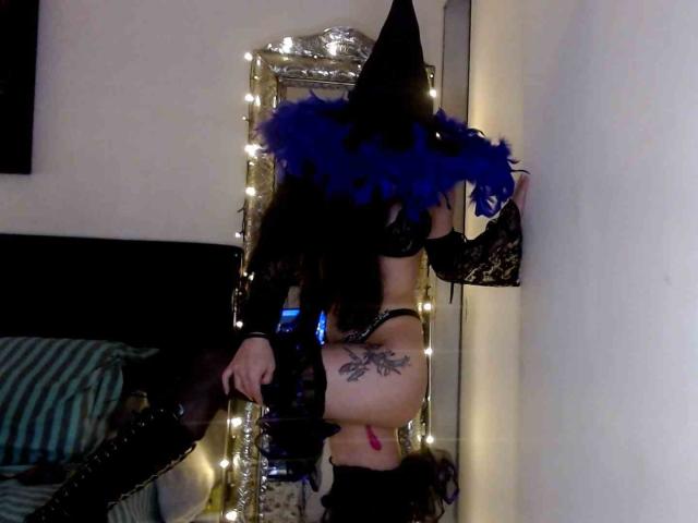 SeleneBrigth in black lace lingerie, blue feathered witch hat, thigh-high boots, and exposed ass, leaning against a mirror with string lights, showing a tattoo on her right thigh.