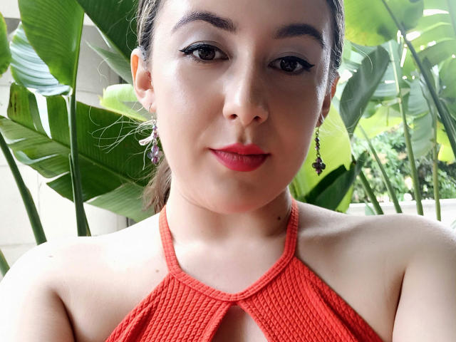MabyLegrand with red lipstick, brown eyes, and red halter top, showing her tits, wearing purple earrings, standing in front of green leaves, smiling slightly.