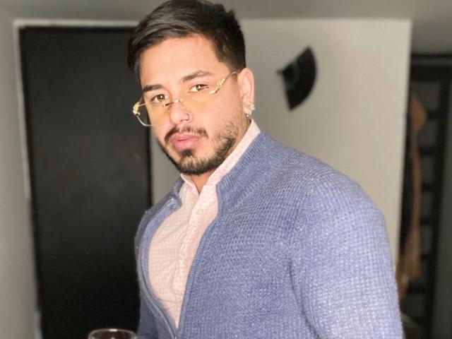 Een persoon met kort donker haar, een baard en een bril, gekleed in een blauwe cardigan over een roze shirt, kijkt naar de camera en heeft een lichte grijns, in een goed verlichte kamer..