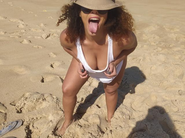 VolupMilf with curly brown hair, tan skin, and big tits, wearing a white tank top, sun hat, and sunglasses, sticking out her tongue, bending over on a sandy beach.