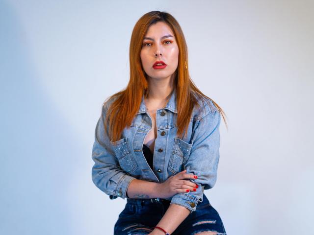 AnnyCraffort, con lunghi capelli rossi, indossa una giacca di jeans blu e pantaloni neri strappati, seduta con le braccia incrociate, mostrando rossetto e smalto rosso, e guarda verso la fotocamera..