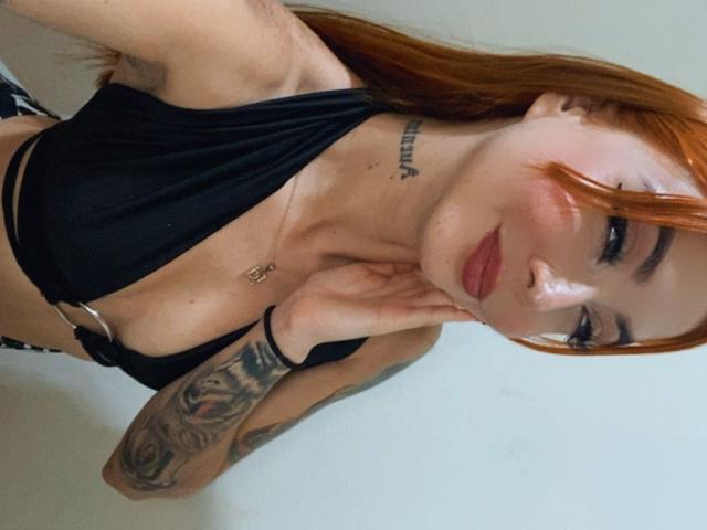 Victoria Gusman, avec des cheveux roux, un haut noir à encolure plongeante, des tatouages sur le bras droit, une main posée sur le menton, un bracelet noir et du rouge à lèvres rose, montrant de petites seins et un sourire discret..