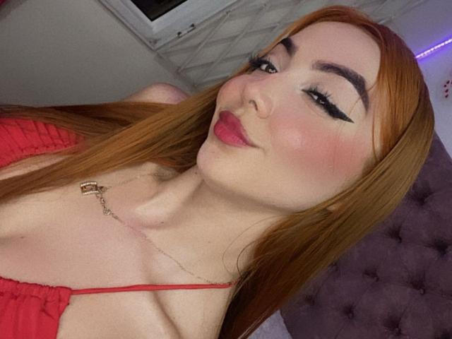VictoriaGusman with red hair, black eyeliner, pink blush, and red lipstick, wearing a red top, showing her tits, lying on a purple bed, smiling at the camera.