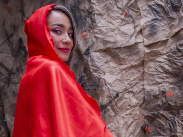 Sarahy Moore, con una capucha roja brillante, lápiz labial rosa y cabello gris, mirando hacia atrás con una sonrisa sutil. Lleva una capa de satén rojo, que muestra ligeramente su trasero..