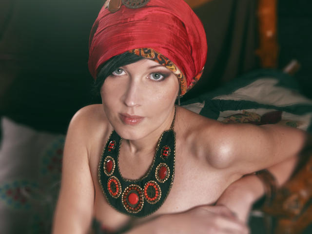 VeraWatts with a red headscarf, black beaded necklace, and bare tits, looking up with green eyes, pink lips, and fair skin, leaning forward on a patterned surface.