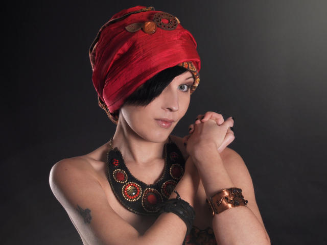 VeraWatts mit einem roten Turban, einer schwarzen Halskette und einem Kupferarmband, die Händchen haltend, mit einer Tätowierung auf ihrer rechten Schulter, kurzen schwarzen Haaren und einem leichten Lächeln..
