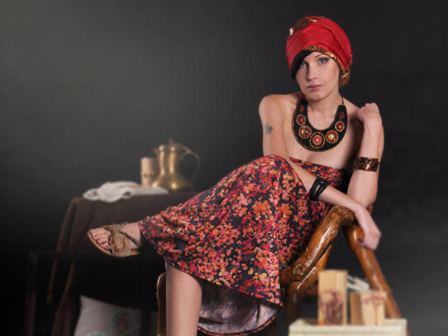 VeraWatts, con i capelli corti e scuri, un foulard rosso, un vestito floreale, una collana nera e un braccialetto, seduta su una sedia di legno, con le gambe incrociate, che mostra i suoi piccoli seni, in una stanza buia..