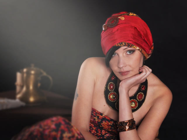 VeraWatts, con un pañuelo rojo en la cabeza, un collar negro con gemas rojas y una pulsera de latón, apoyando el mentón en la mano, mirando a la cámara, mostrando sus pequeños senos..