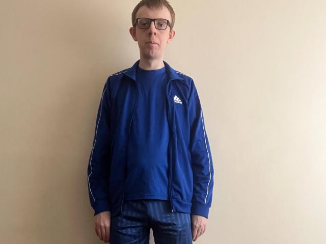 Liam Miller, con gli occhiali, giubbotto blu Adidas, camicia blu e jeans blu, in piedi, che guarda leggermente verso l'alto, capelli castani corti, espressione neutra, parete beige sullo sfondo..