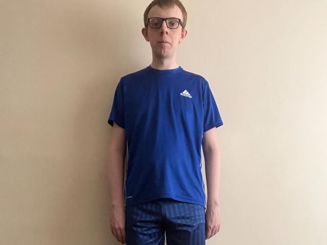 Liam Miller staat in een blauw Adidas T-shirt en donkerblauwe gestreepte broek, draagt een zwarte bril en heeft een neutrale uitdrukking, voor een effen beige muur..