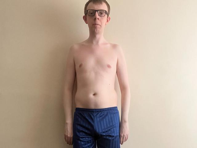 LiamMiller, shirtless with glasses, wearing blue shorts, standing against a plain wall. He has a slim build, small nipples, and a slightly visible belly button.