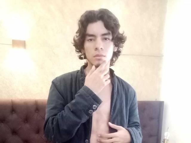 DylanWyster with curly dark hair, wearing an open black shirt, touching his chin with one hand and his chest with the other, looking serious.
