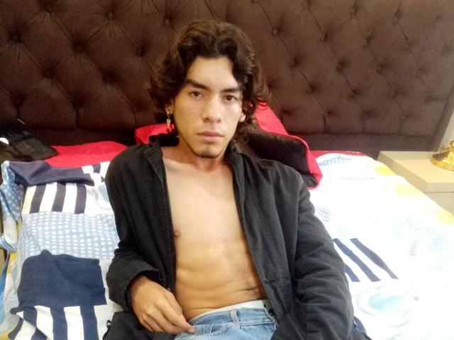 DylanWyster with curly brown hair, shirtless, black robe open, sitting on bed, showing a toned, tan-skinned torso, and a slight tattoo on right side, looking serious.