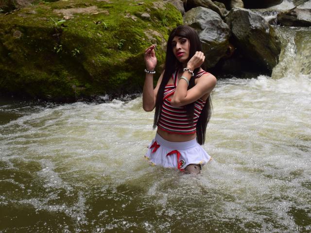 Catalina Castro se tient à mi-mollet dans une rivière, vêtue d'un débardeur rayé et d'une mini-jupe blanche avec des nœuds rouges, avec de longs cheveux bruns et des bijoux, et elle regarde l'appareil photo..