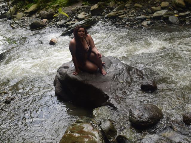NaomiQuinn está sentada sobre una roca en un río caudaloso, vistiendo un bikini negro, con el cabello castaño mojado, sonriendo a la cámara, mostrando su trasero y sus pequeños senos..